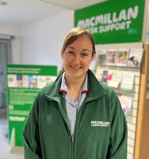 Photo of Emma James, a young white female, wearing a green macmillan cancer support fleece in a hospital information area