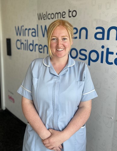 Photo of Jamie-Lee Hayes wearing a light blue maternity support worker uniform standing in front of a sign saying Wirral Women and Children's Hospital
