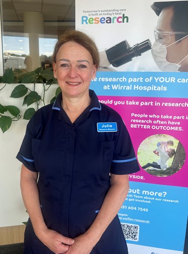 Photo of Julie Grindey Research Midwife wearing a navy tunic with light blue piping standing in front of a tall banner with images and text including Make research part of your care at Wirral Hospitals