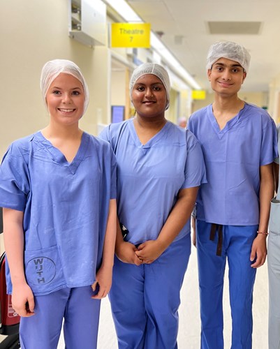 Three A level students wearing blue scrubs and theatre caps standing in a corridor with a sign saying Theatre 7